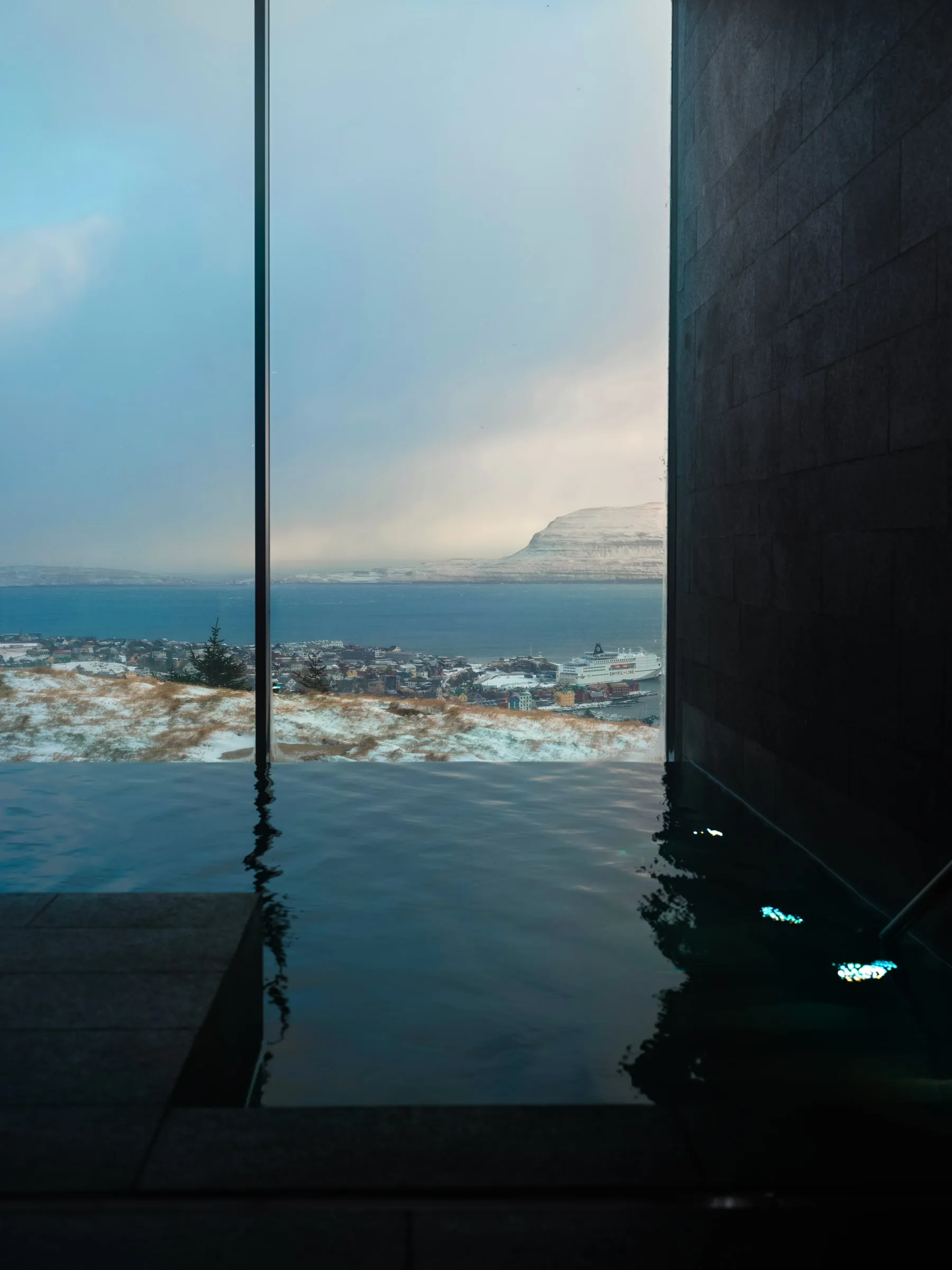 The pool meeting the glass wall with the town below, and the dark water of the indoor pool