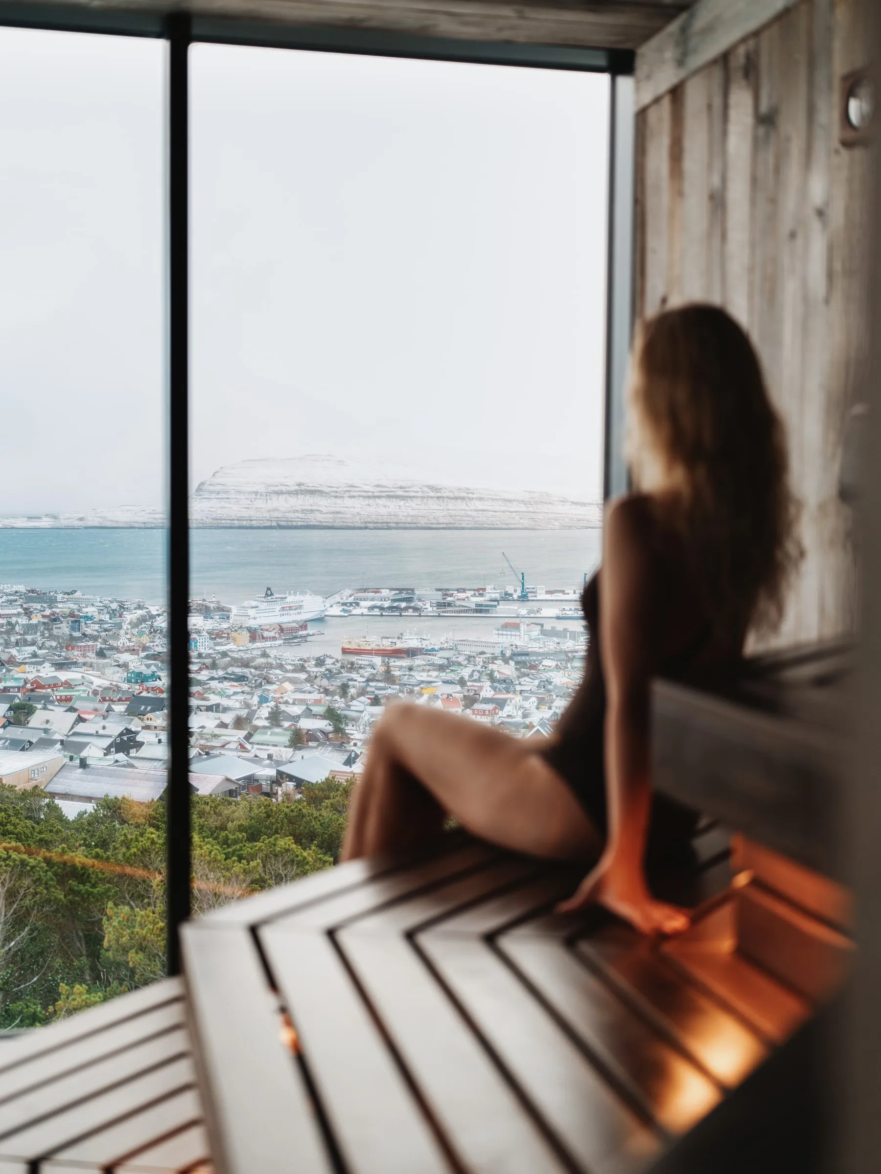 A bather on the sauna bench by the window, the snow-covered town below