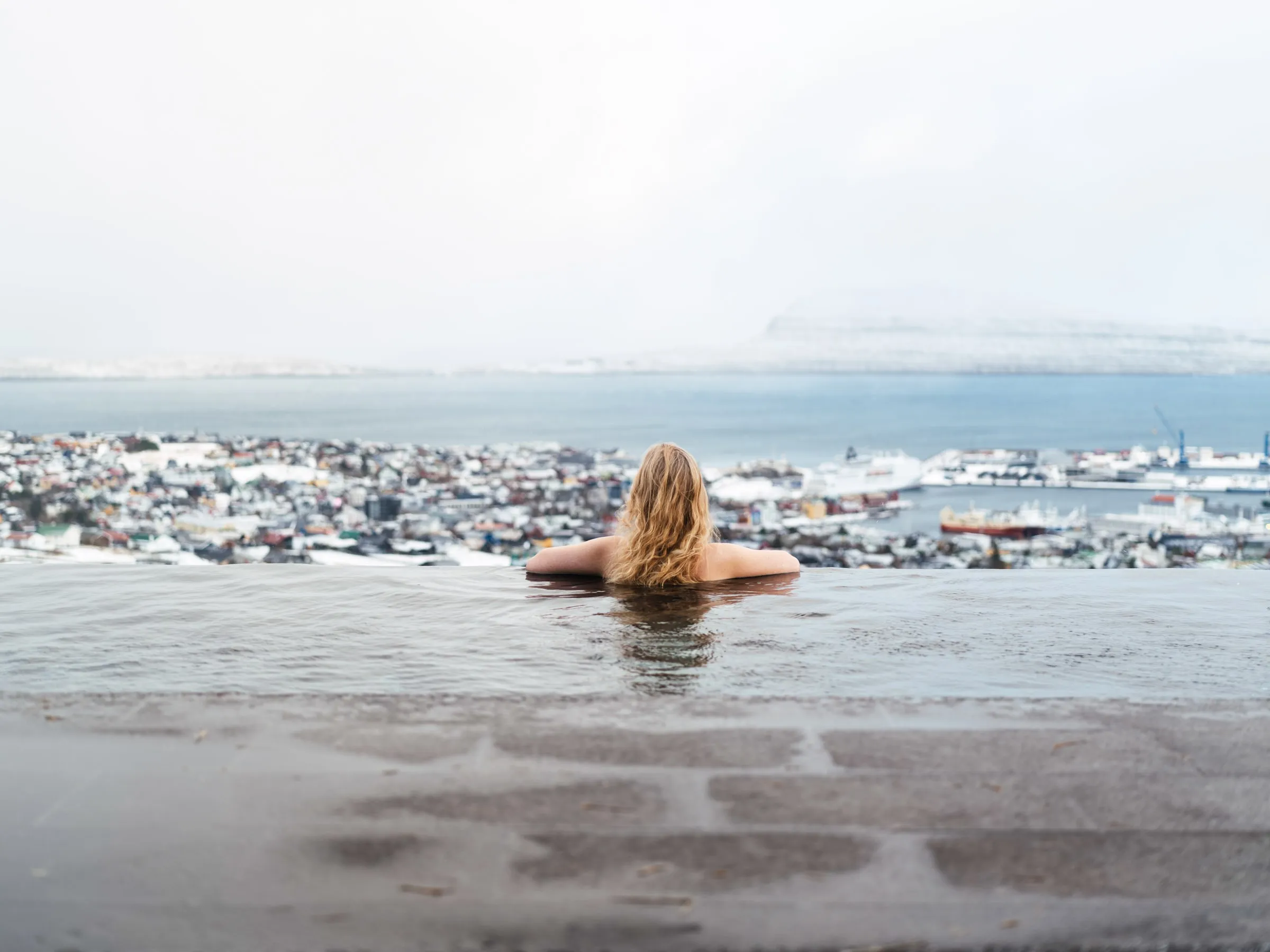 A bather at the edge of the infinity pool, Tórshavn and the snow-covered hills beyond